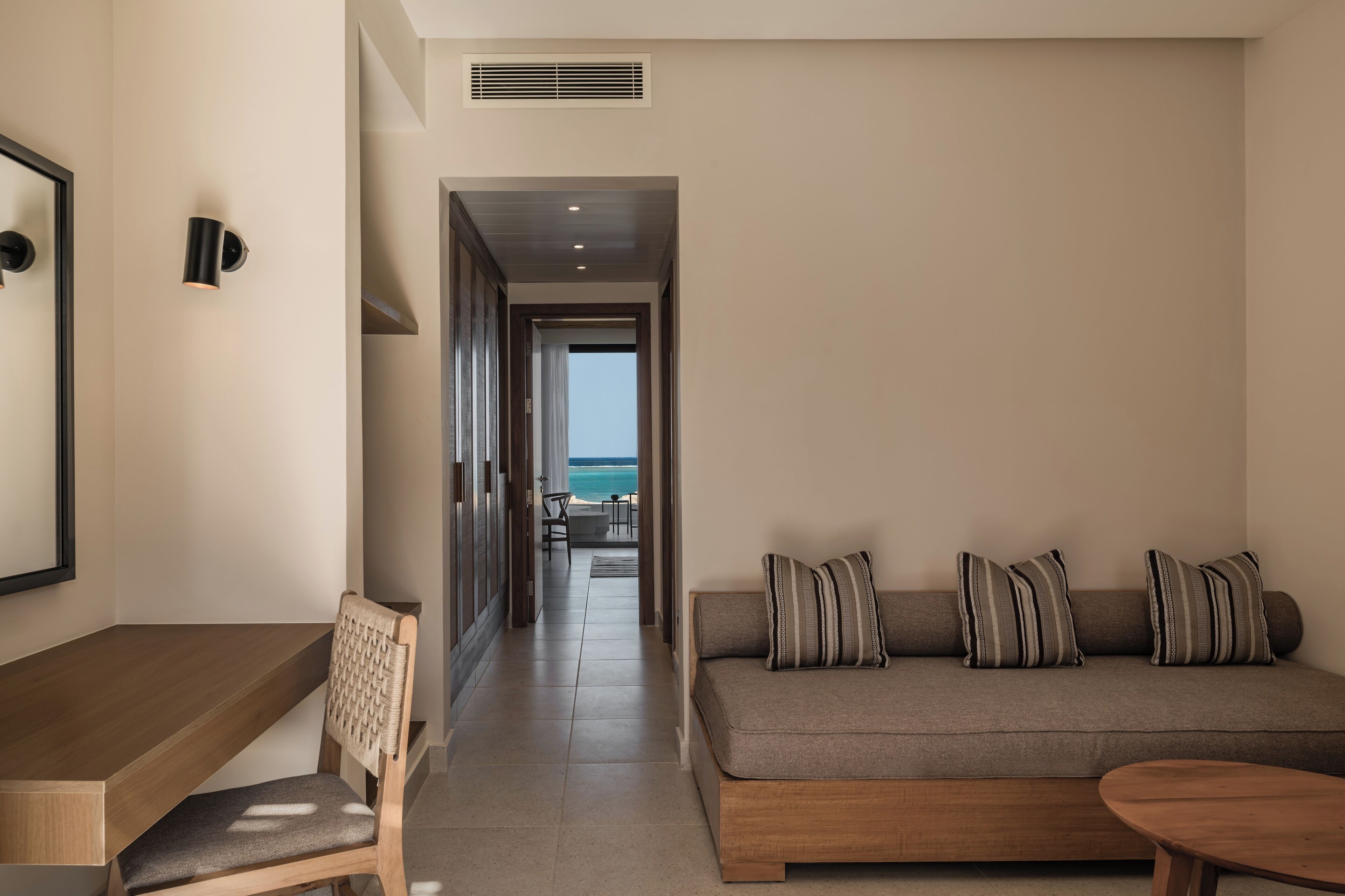 Una habitación de hotel moderna con un escritorio de madera, una silla acolchada, un sofá con almohadones a rayas y vistas al océano a través de un vano abierto.