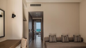 Una habitación de hotel moderna con un escritorio de madera, una silla acolchada, un sofá con almohadones a rayas y vistas al océano a través de un vano abierto.