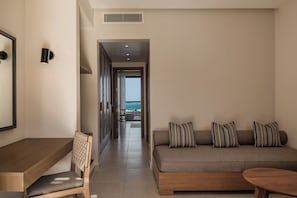 Een moderne hotelkamer met een houten bureau, een comfortabele fauteuil, een bank met gestreepte kussens en uitzicht op de oceaan door een open deur.