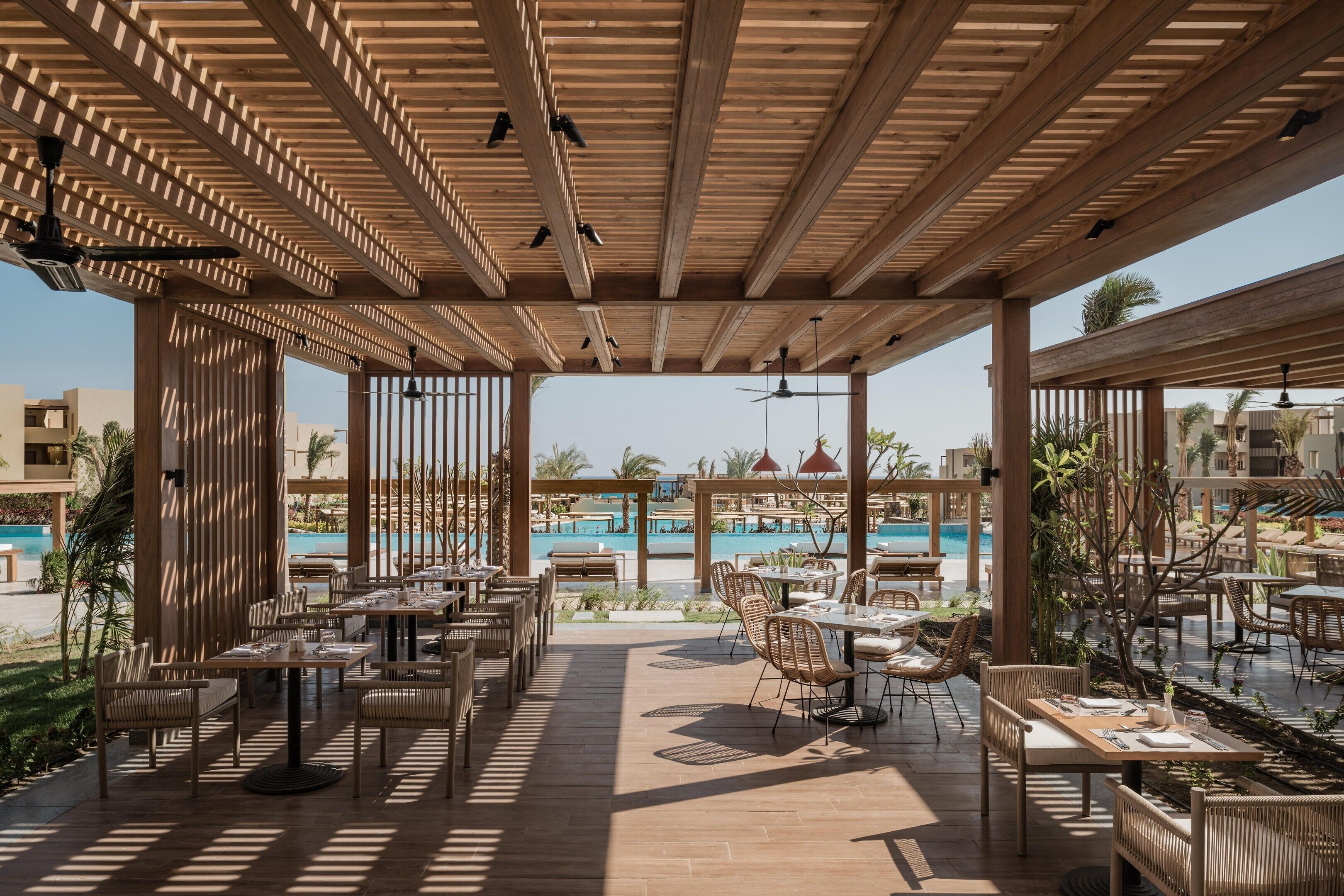Een terras met houten pergola’s, tafels en stoelen met uitzicht op een zwembad.