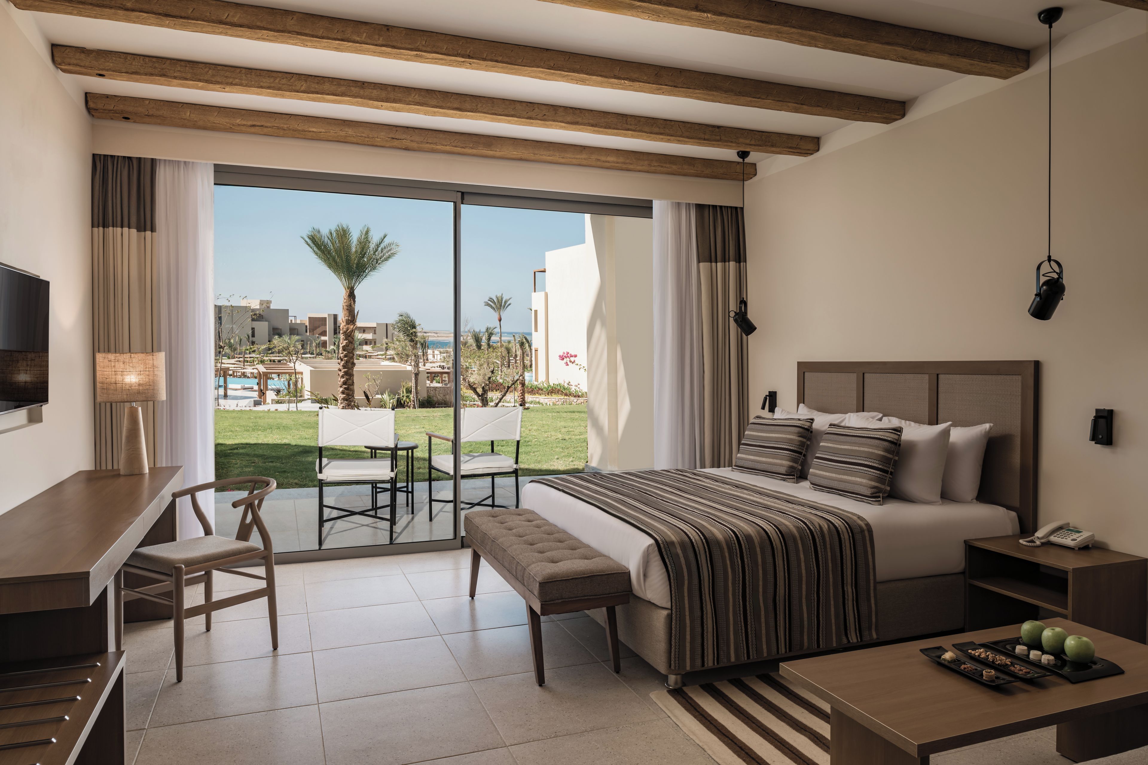 A hotel room with a bed, bedside table, a desk with a chair, a TV, and a view of a pool and palm trees.