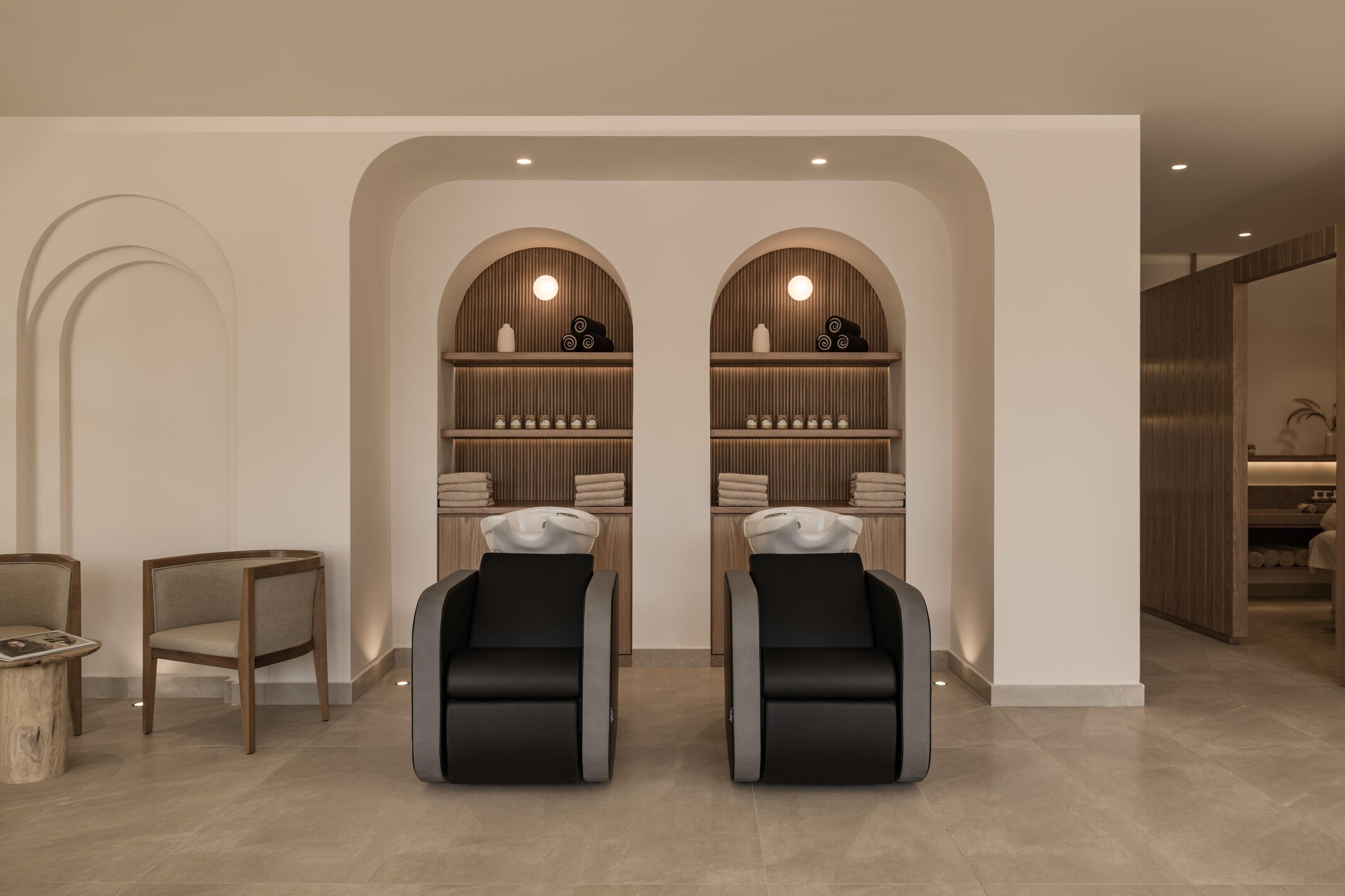A modern spa room with two massage chairs, a wooden vanity with a sink, and a shelf with towels and candles.