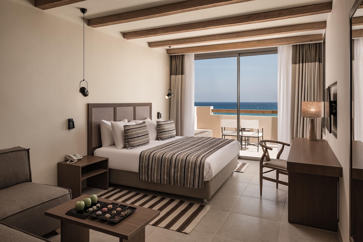 Habitación de hotel con cama, mesitas de noche, escritorio, silla, una pequeña mesa con aperitivos y vista al mar.