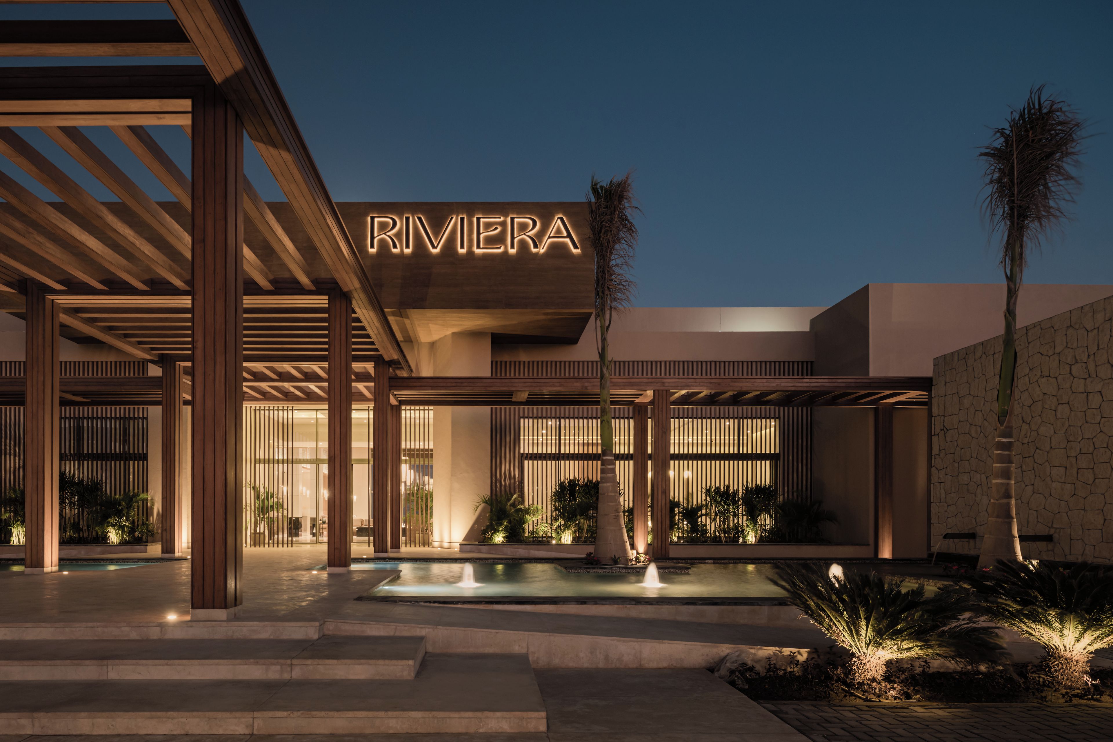 Un edificio moderno con el nombre "RIVIERA" iluminado en la fachada, que cuenta con una piscina y un área de estar al aire libre.