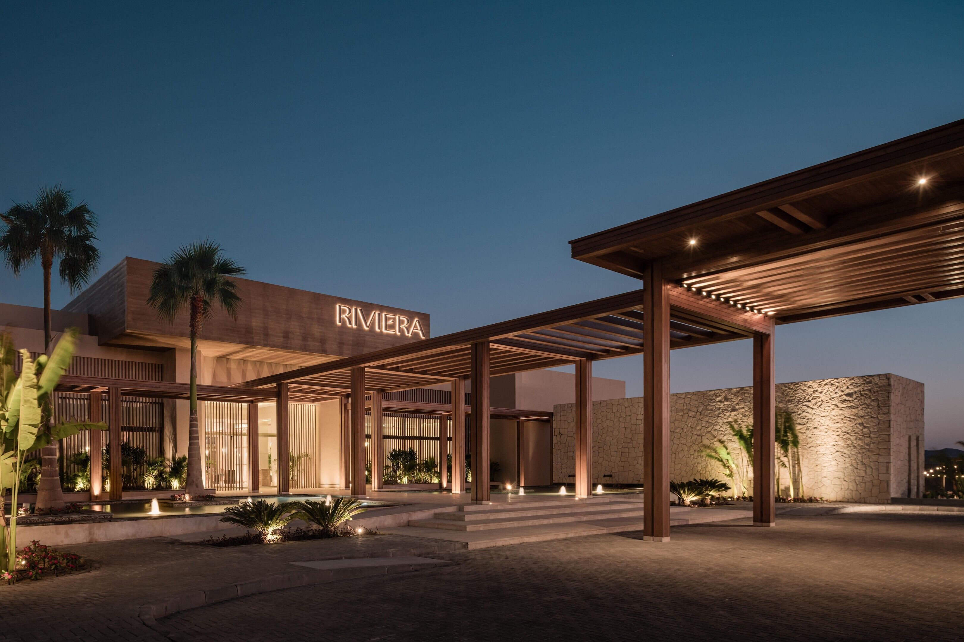 Una entrada de hotel moderna con un paseo techado, palmeras y un área de piscina.