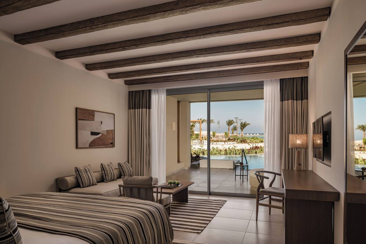 A modern hotel room with a large bed, a sofa, a desk, and a view of a pool and palm trees.