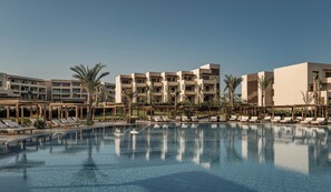 A resort pool with lounge chairs and palm trees, surrounded by multi-story buildings with balconies.