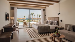 A hotel room with a bed, a desk, a sofa, and a dining table. There is a large window with a view of palm trees and a pool. - JAZ Elite Riviera (Marsa Alam)