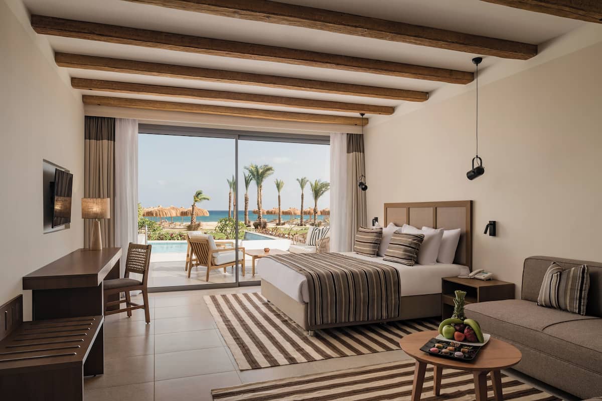 A hotel room with a bed, a desk, a sofa, and a dining table. There is a large window with a view of palm trees and a pool.