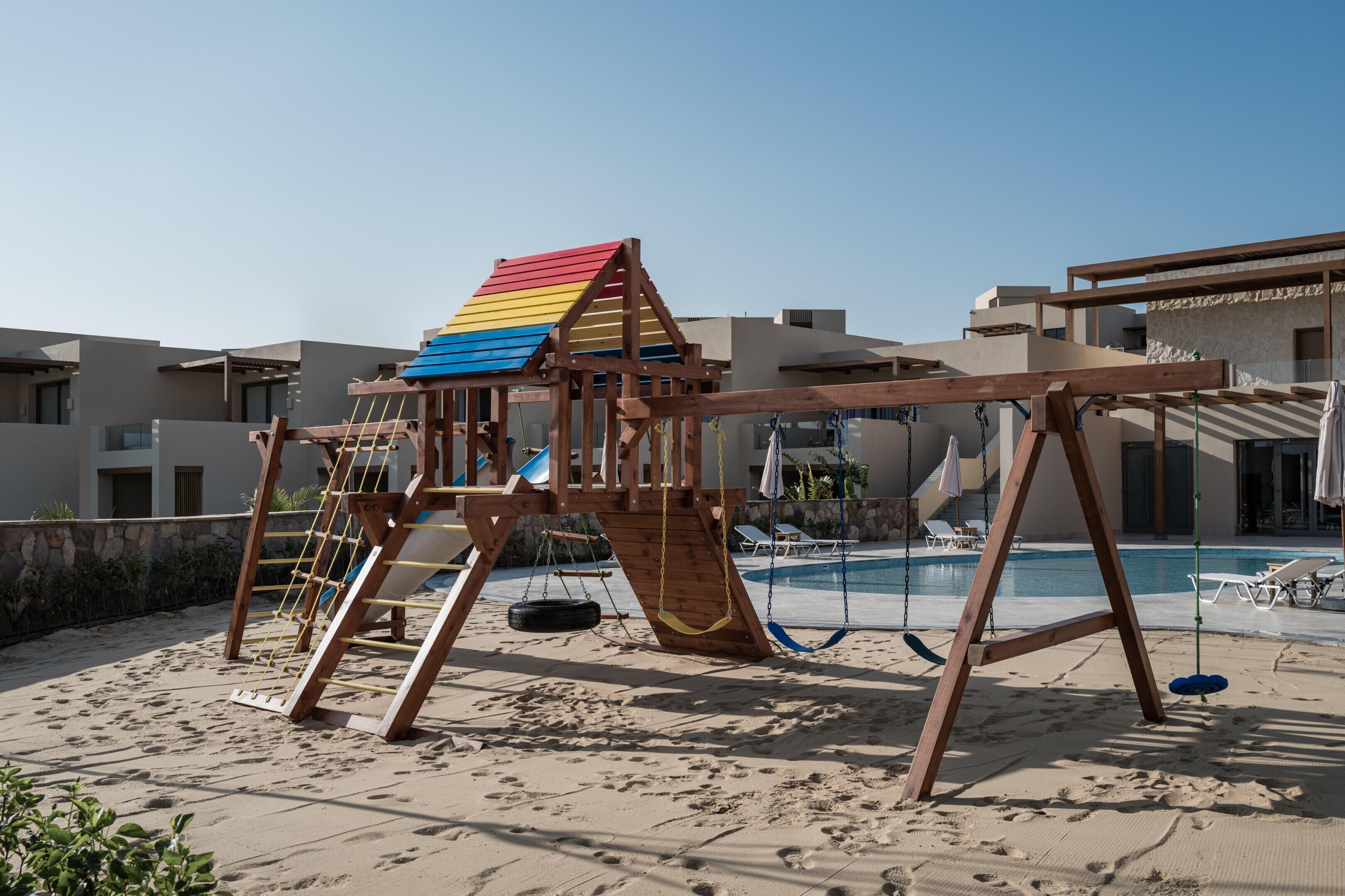 A playground with a swing set and a slide, located on a sandy area with a pool and lounge chairs in the background.