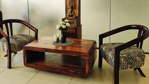A wooden coffee table with a vase of flowers and a Buddha statue, flanked by two patterned chairs in a room with a plain wall and tiled floor. - La Cozy Residency (Gurugram)