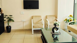 A modern waiting area with two beige chairs, a glass coffee table, a potted plant, and a flat-screen TV mounted on the wall. - La Cozy Residency (Gurugram)