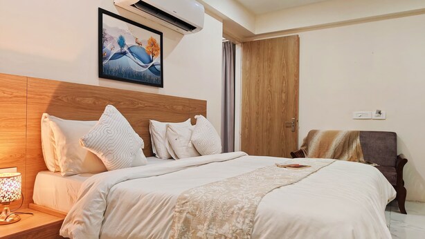 A hotel room with a wooden headboard, a bed with white bedding and patterned pillows, a bedside table with a lamp, and a painting of a landscape on the wall. - La Cozy Residency (Gurugram)