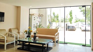 A reception area with a sofa, chairs, a coffee table with flowers, a TV, and a view of parked cars through large windows. - La Cozy Residency (Gurugram)