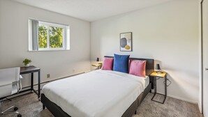 Un dormitorio con una cama, un escritorio, una silla, un ventanal y una mesita de noche con una lámpara.
