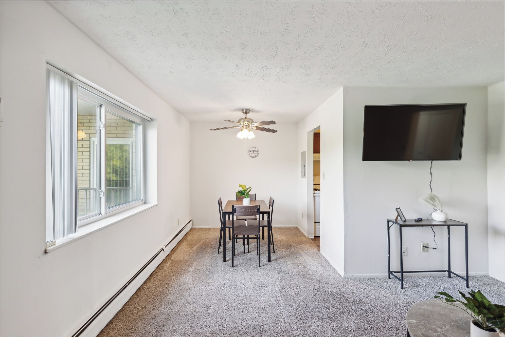 A dining area with a table and chairs, a ceiling fan, a TV on the wall, and a small side table with a plant.