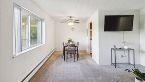 Un coin repas comprenant une table et des chaises, un ventilateur de plafond, un téléviseur fixé au mur et une petite table d’appoint avec une plante.