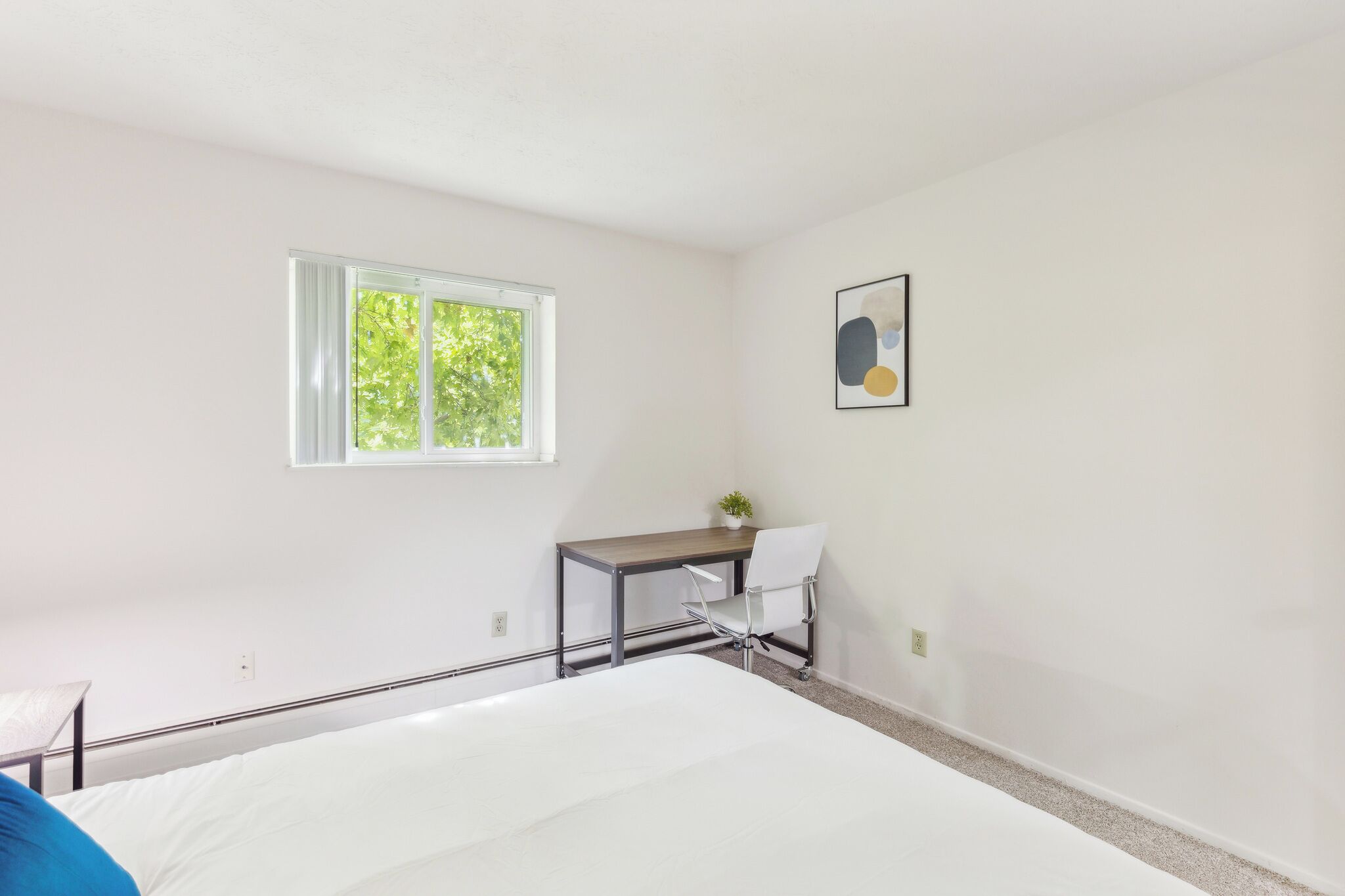 Een slaapkamer met een bed, een bureau met stoel, een raam en een aan de muur hangend schilderij.