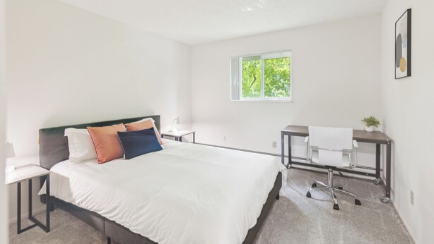 A modern hotel room with a bed, a desk, and a chair. There is a window with a view of trees.