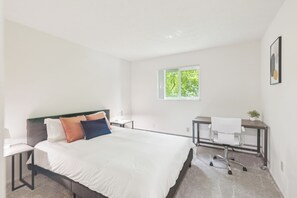A modern hotel room with a bed, a desk, and a chair. There is a window with a view of trees.