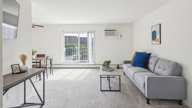 Una sala de estar moderna con un sofá gris, una mesa de centro, una mesa de comedor con sillas, un televisor montado en la pared y un amplio ventanal con vistas a edificios.