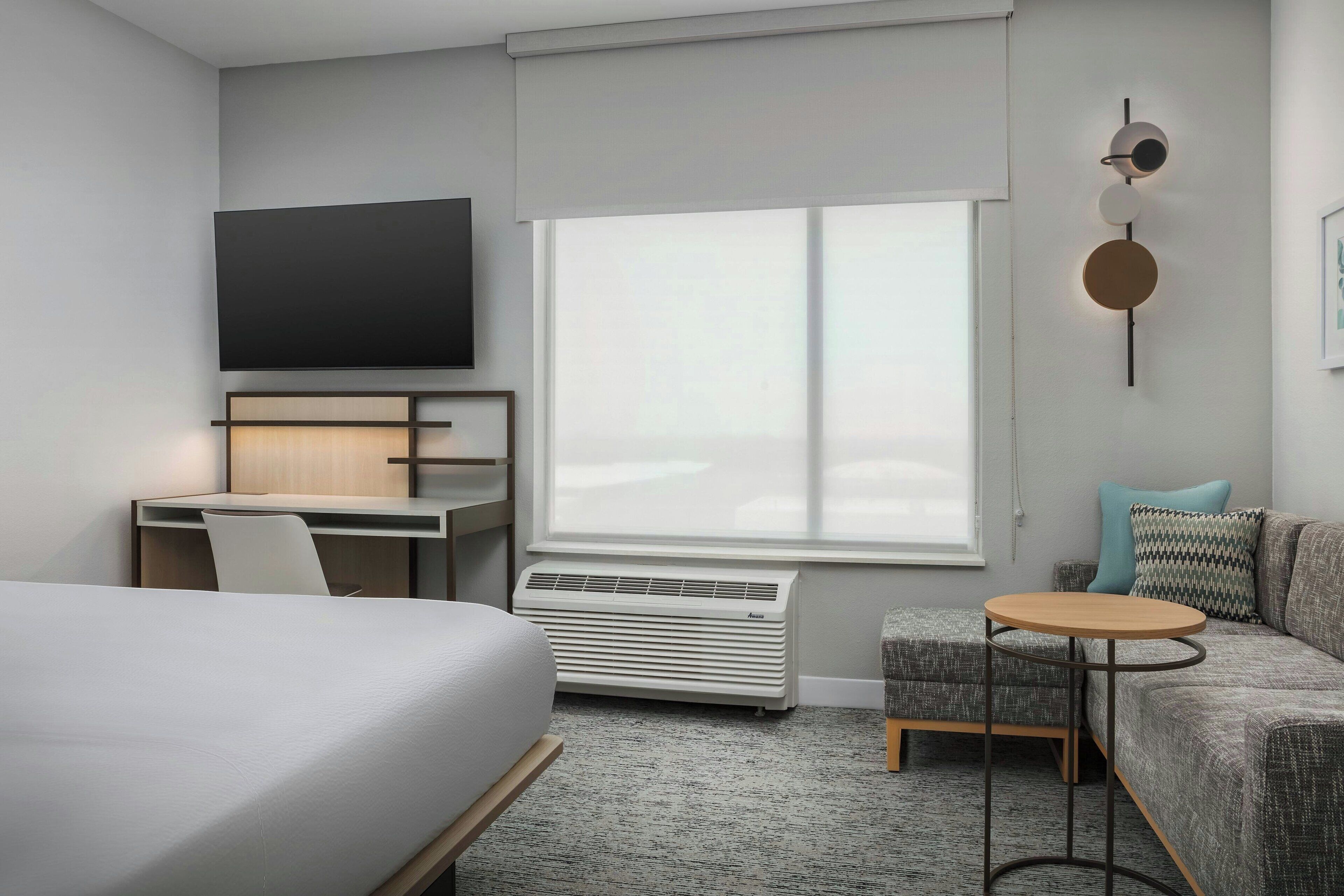 A modern hotel room with a bed, a desk with a chair, a small round table, a sofa, a TV mounted on the wall, and a large window.