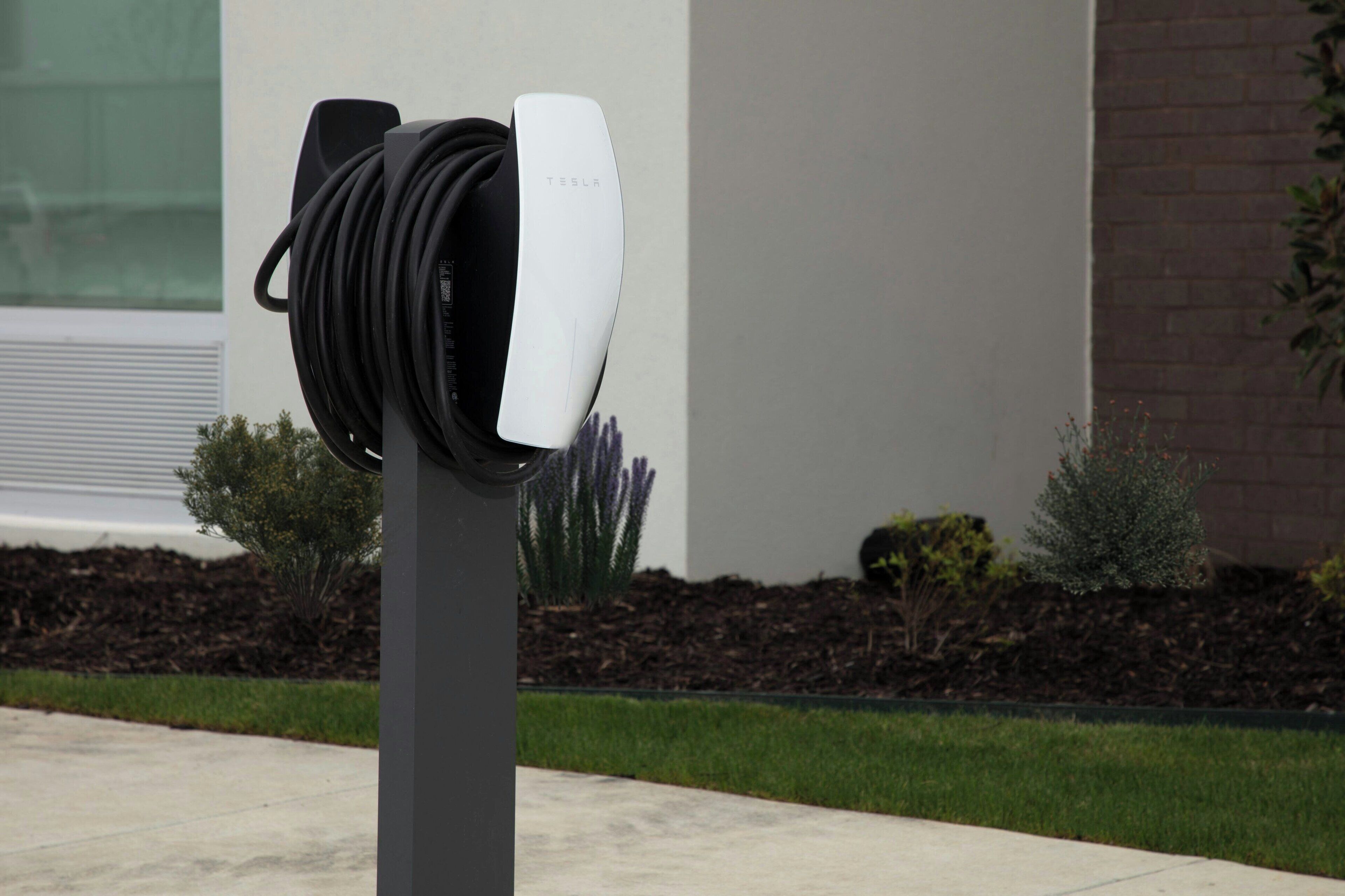 A Tesla charging station with a white and black charging unit, set against a building with a brick wall and a landscaped garden.