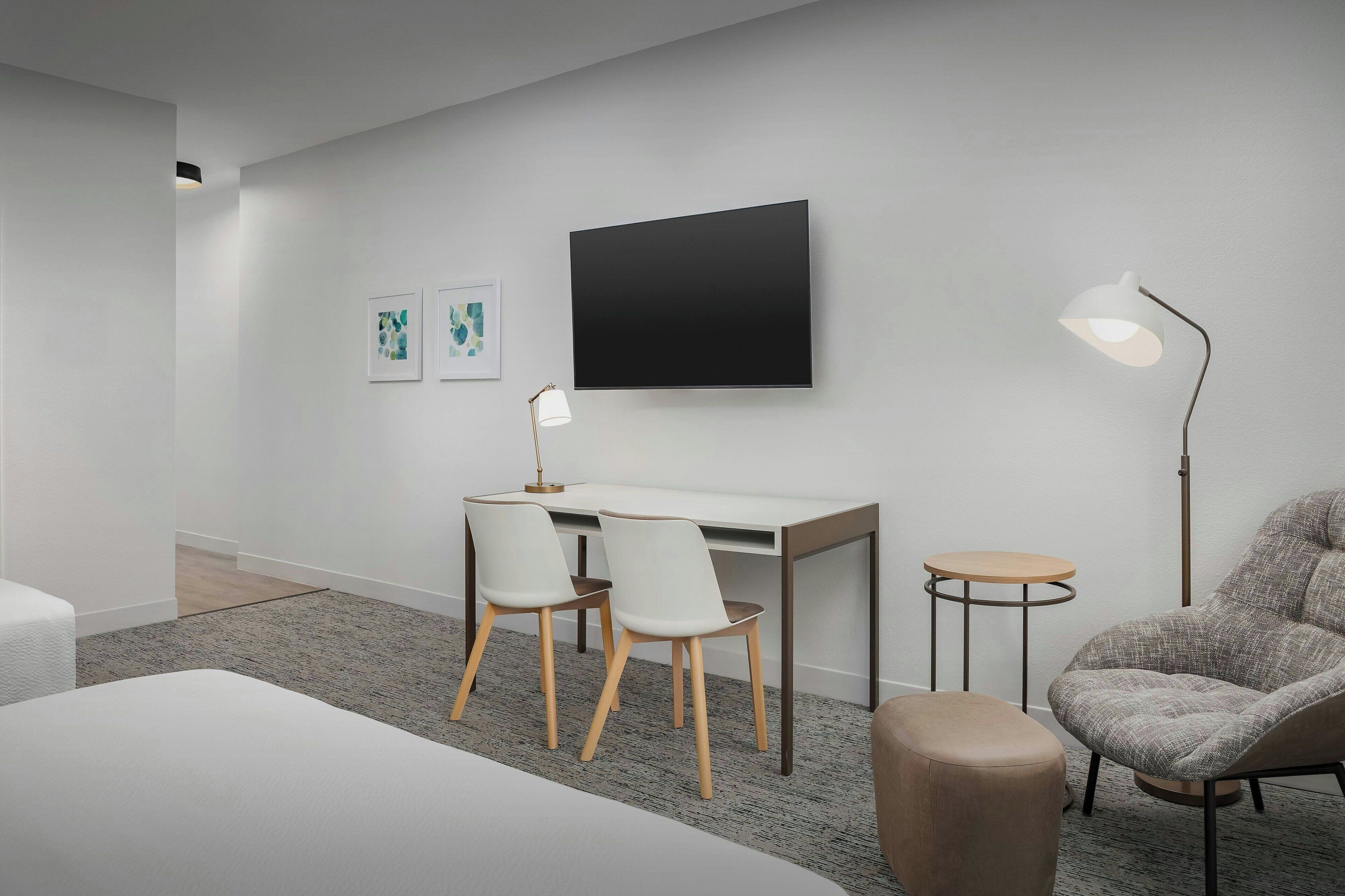 A modern hotel room with a white desk, two chairs, a large flat-screen TV, a floor lamp, and a grey armchair.