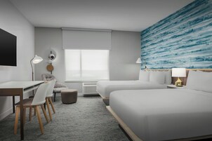 A modern hotel room with a large bed, a desk with chairs, a flat-screen TV, and a large window with a view of the ocean.