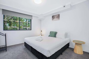A modern bedroom with a large window, a bed with white bedding, a green pillow, and a small wooden side table.