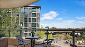 Un balcón con dos sillas y una mesa, con vista a un edificio moderno y una calle concurrida.