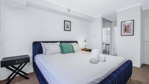 Una cama bien hecha con edredón blanco y azul, una mesita de noche con lámpara y un cuadro en la pared.