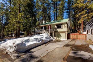 A two-story cabin with a green roof, surrounded by tall pine trees and a snow-covered ground.