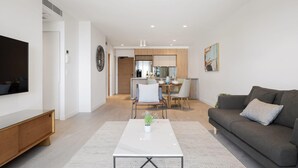 A modern living room with a sofa, a coffee table, and a TV. There is a dining area with a table and chairs.
