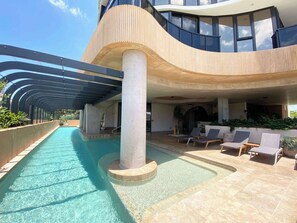 A swimming pool with a curved roof structure, lounge chairs, and a modern building with large windows.