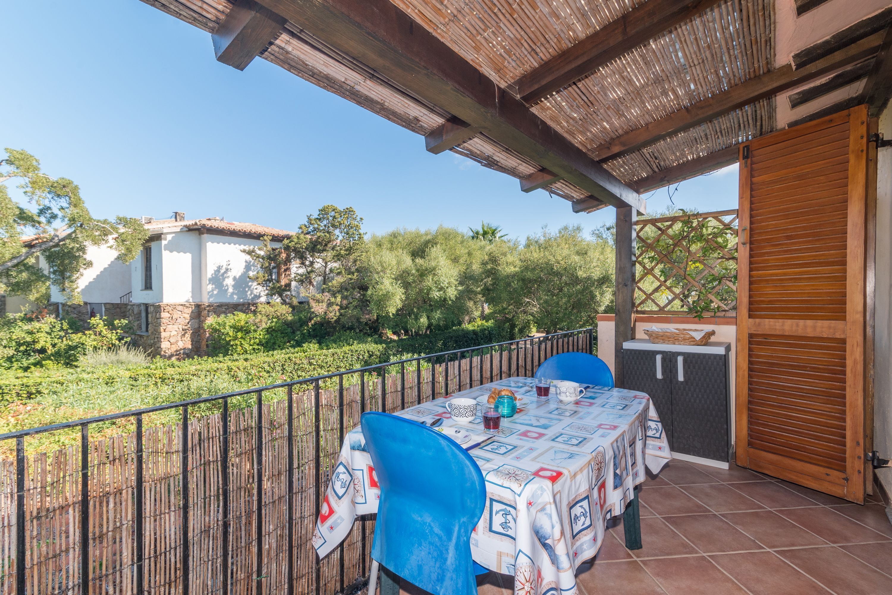 Um pátio externo com mesa e cadeiras, uma pérgula de madeira e vista para o jardim e edifícios ao redor.