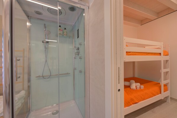 Una habitación con cabina de ducha de vidrio, una cama litera con ropa de cama naranja y una puerta blanca.