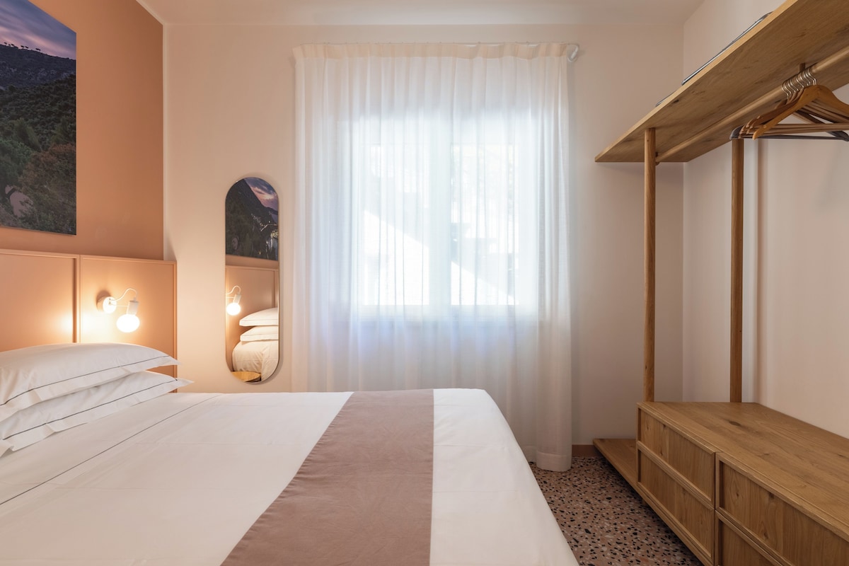 A hotel room with a bed, a wooden bench, a mirror, and a window with sheer curtains.