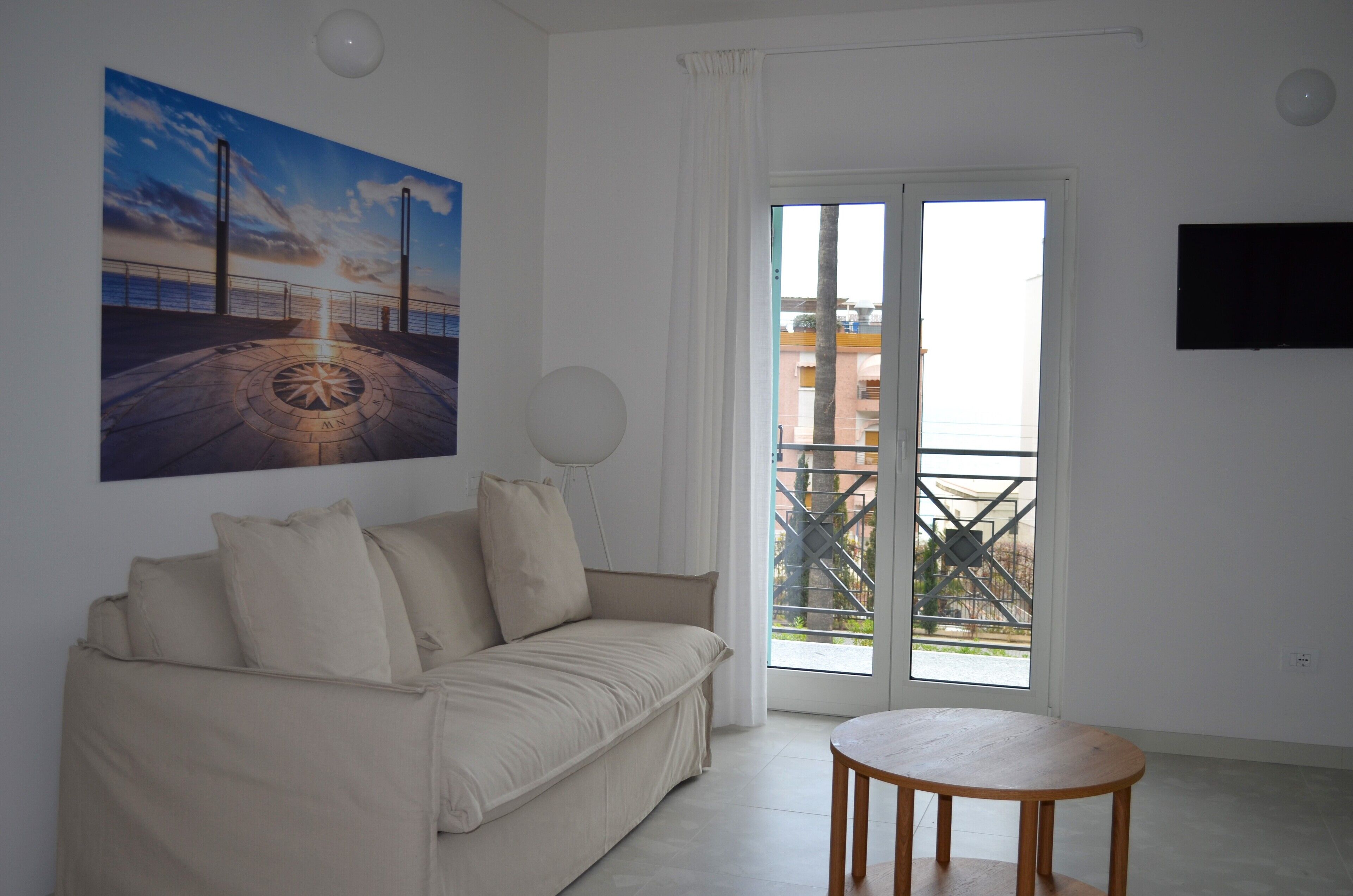 A living room with a beige sofa, a round wooden table, a painting of a sunset, and a balcony with a view of buildings.