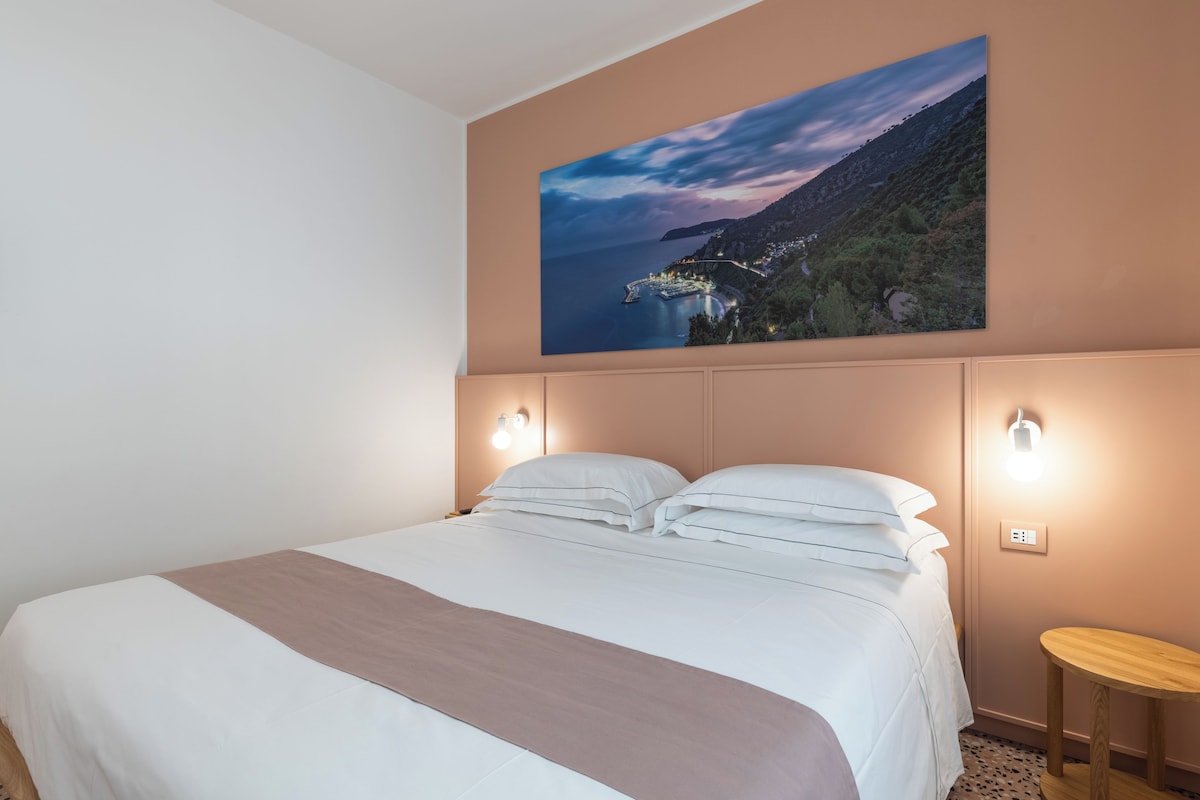 A bedroom with a large bed, a nightstand, a wall-mounted light, and a painting of a coastal landscape.