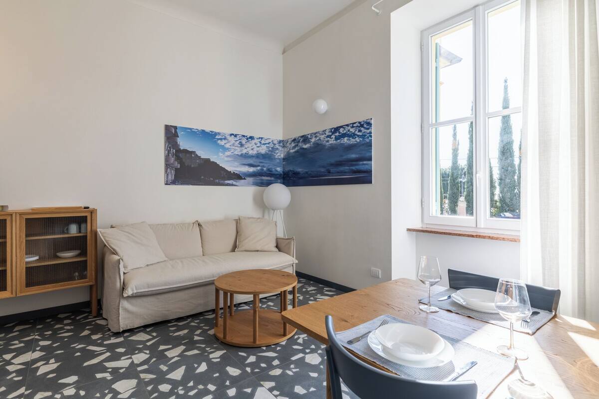 Una sala de estar moderna con un sofá, una mesa de comedor para dos personas, un mueble con vajilla y un ventanal con vistas a árboles.
