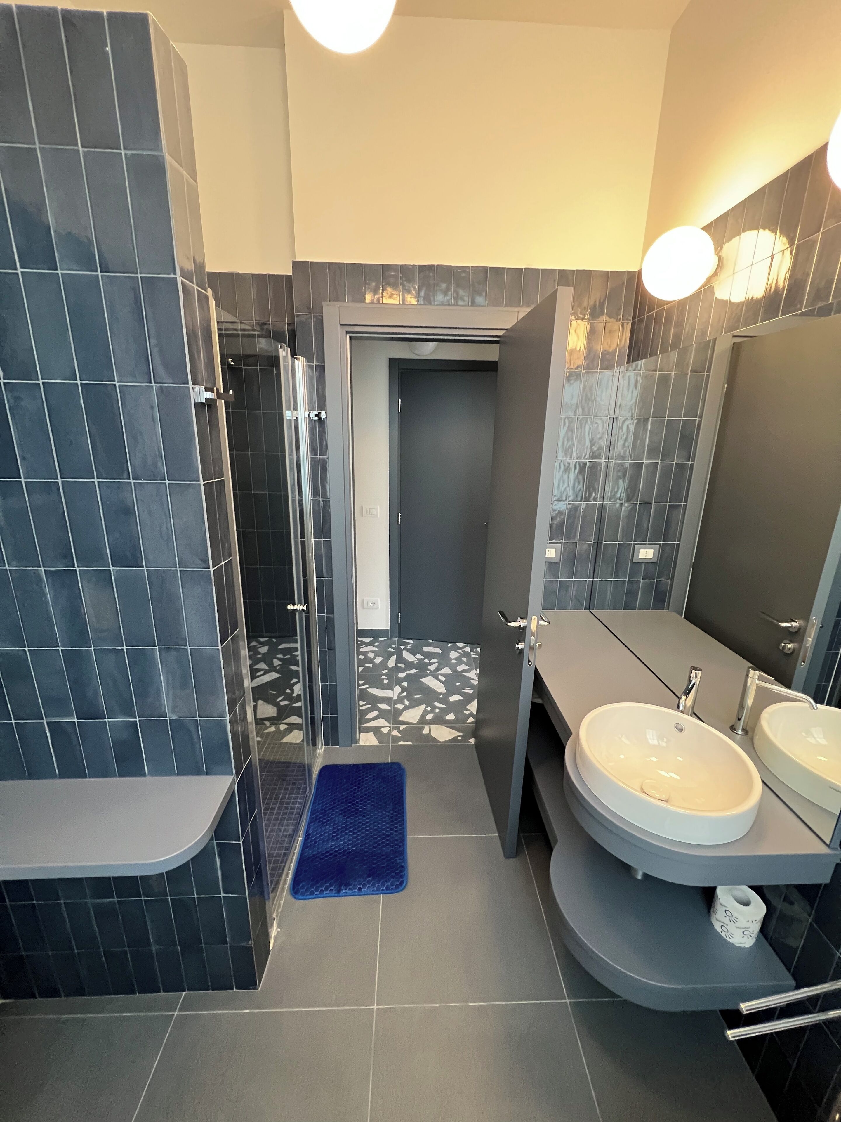 A modern bathroom with dark tiles, a shower area, a sink with a mirror, and a blue mat.