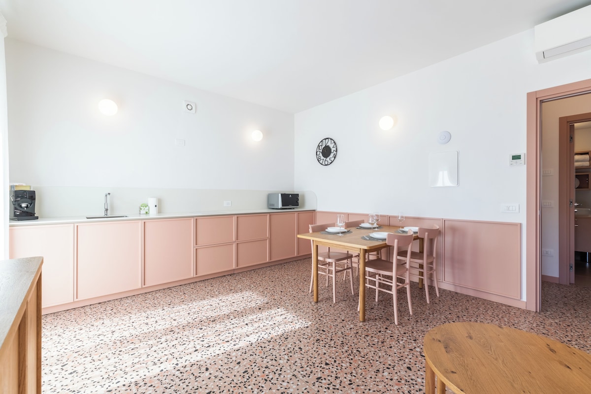 Une cuisine moderne avec un coin repas, dotée d’un sol à motifs, de placards roses et d’une table en bois dressée pour quatre personnes.