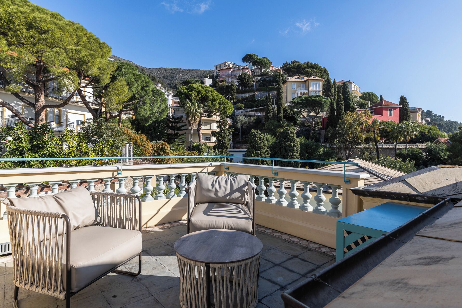 A terrace with outdoor seating, a view of a hillside with buildings, and a railing.