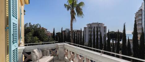 A balcony with a view of the sea, a palm tree, and modern buildings.