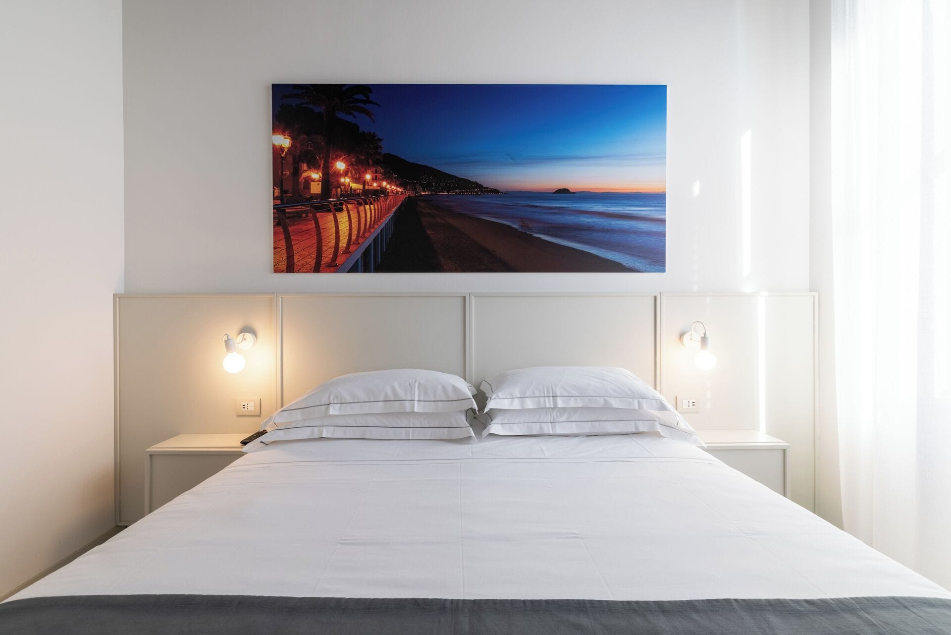 Une chambre d’hôtel avec un grand lit, deux tables de chevet et un tableau représentant une plage au coucher du soleil, accroché au mur.