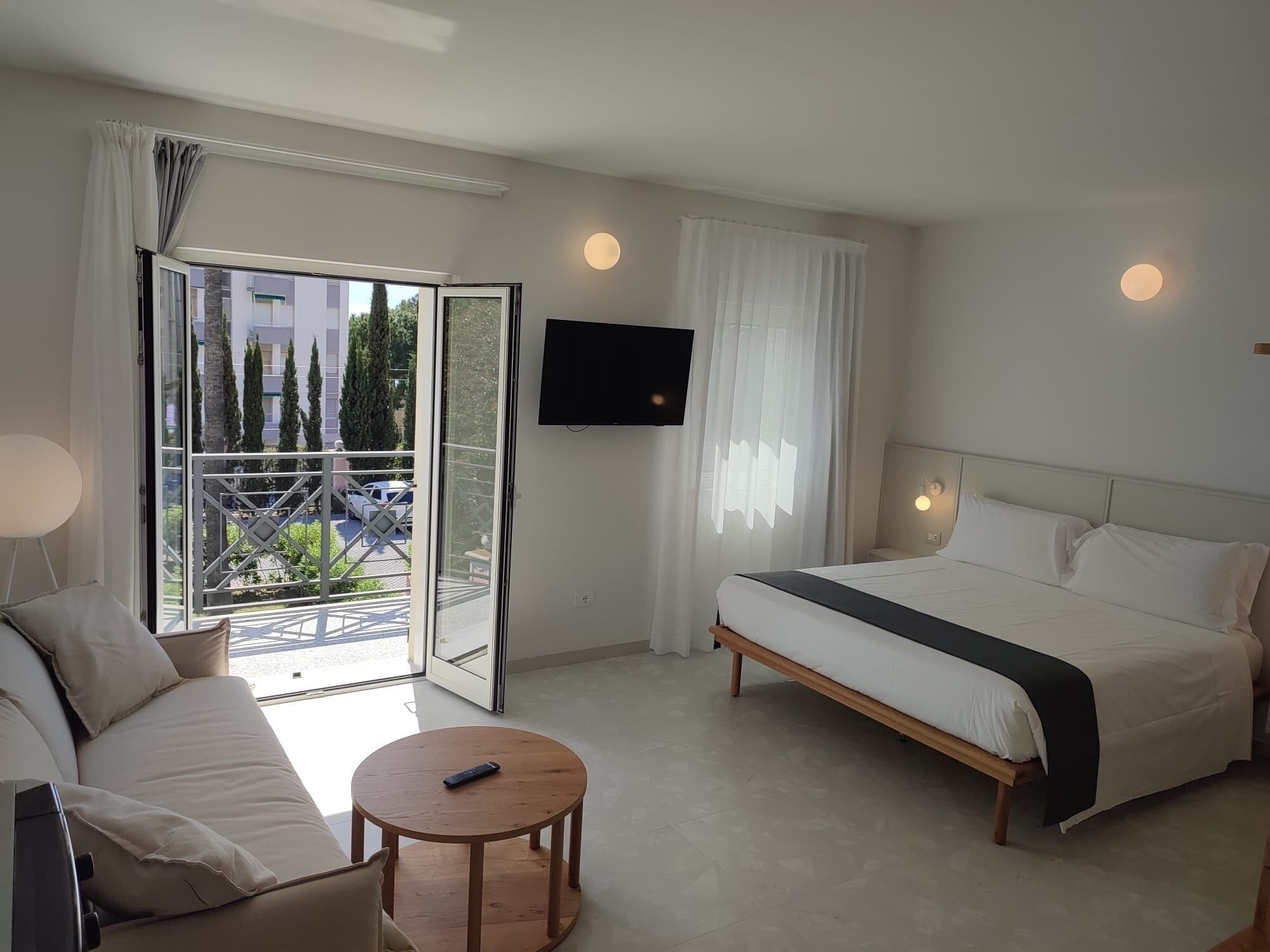 A hotel room with a bed, a sofa, a TV, and a balcony with a view of trees and buildings.