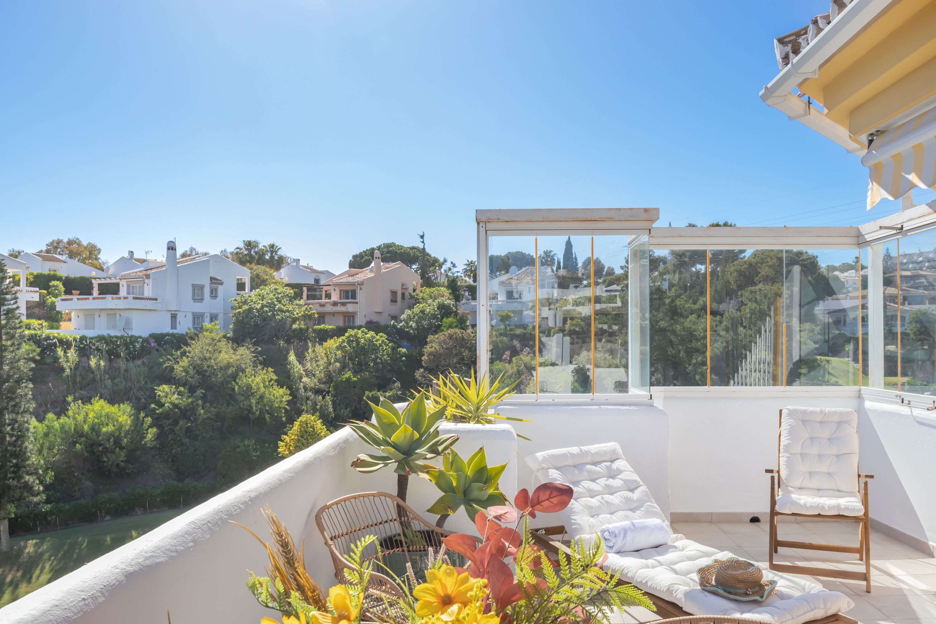A sunlit balcony with a white cushioned sofa, a wicker chair, and a glass enclosure.