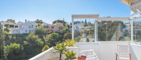 A sunlit balcony with a white cushioned sofa, a wicker chair, and a glass enclosure.