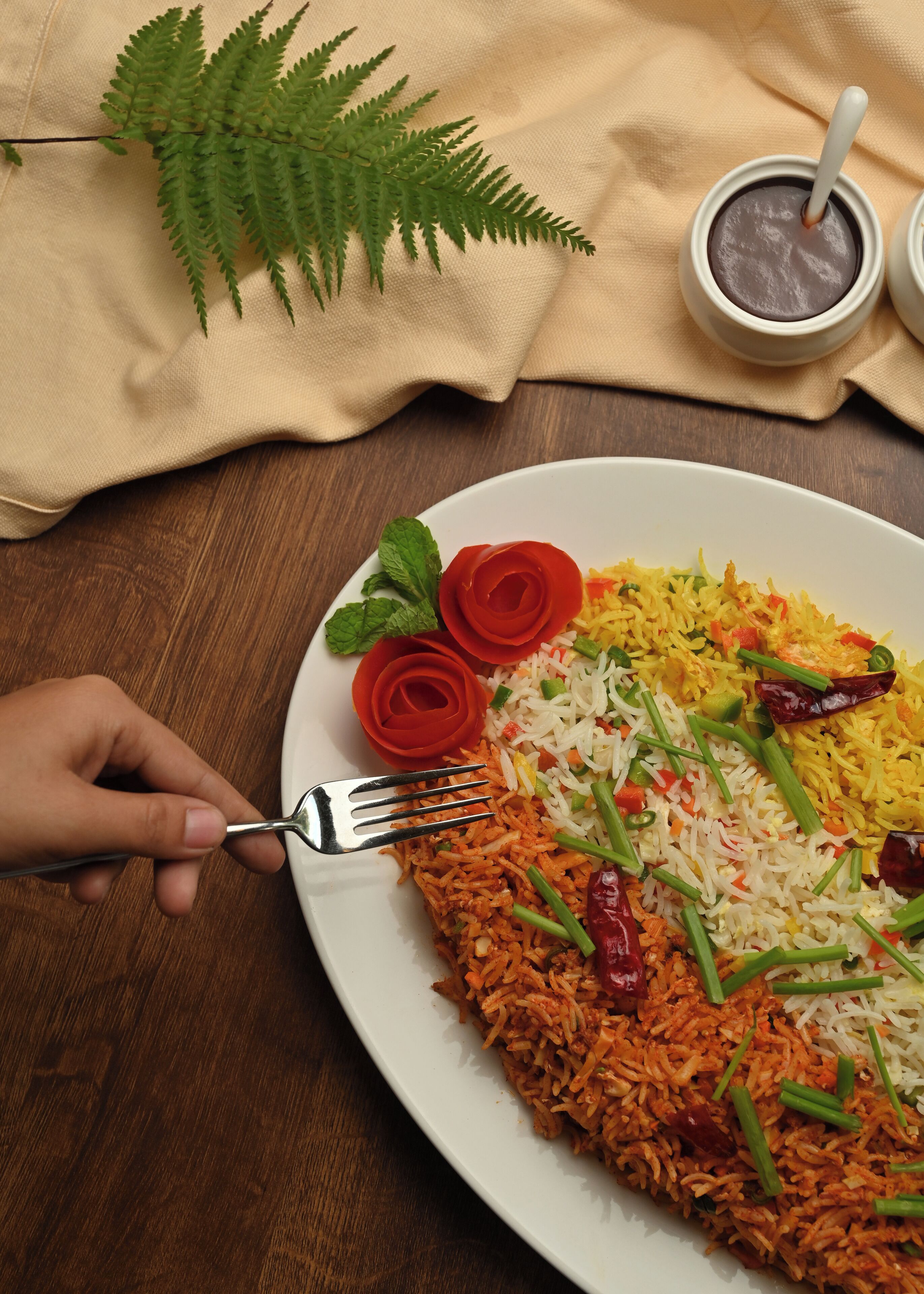 Une assiette de biryani, accompagnée d’une fourchette, décorée de pétales de rose et d’oignons verts, posée sur une table en bois recouverte d’une nappe, avec une feuille de fougère à proximité.